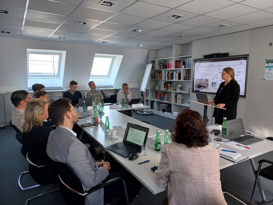 Photo in a meeting room where a workshop is being held with around 10 people. They are sitting in a U-shape on tables and listening to the speaker's PowerPoint presentation.